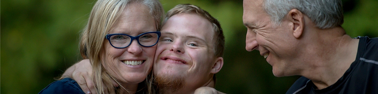 A young man with Down syndrome smiles warmly between his parents, outdoors.