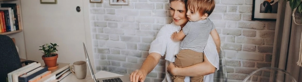 A woman holding a toddler works on a laptop at a desk.